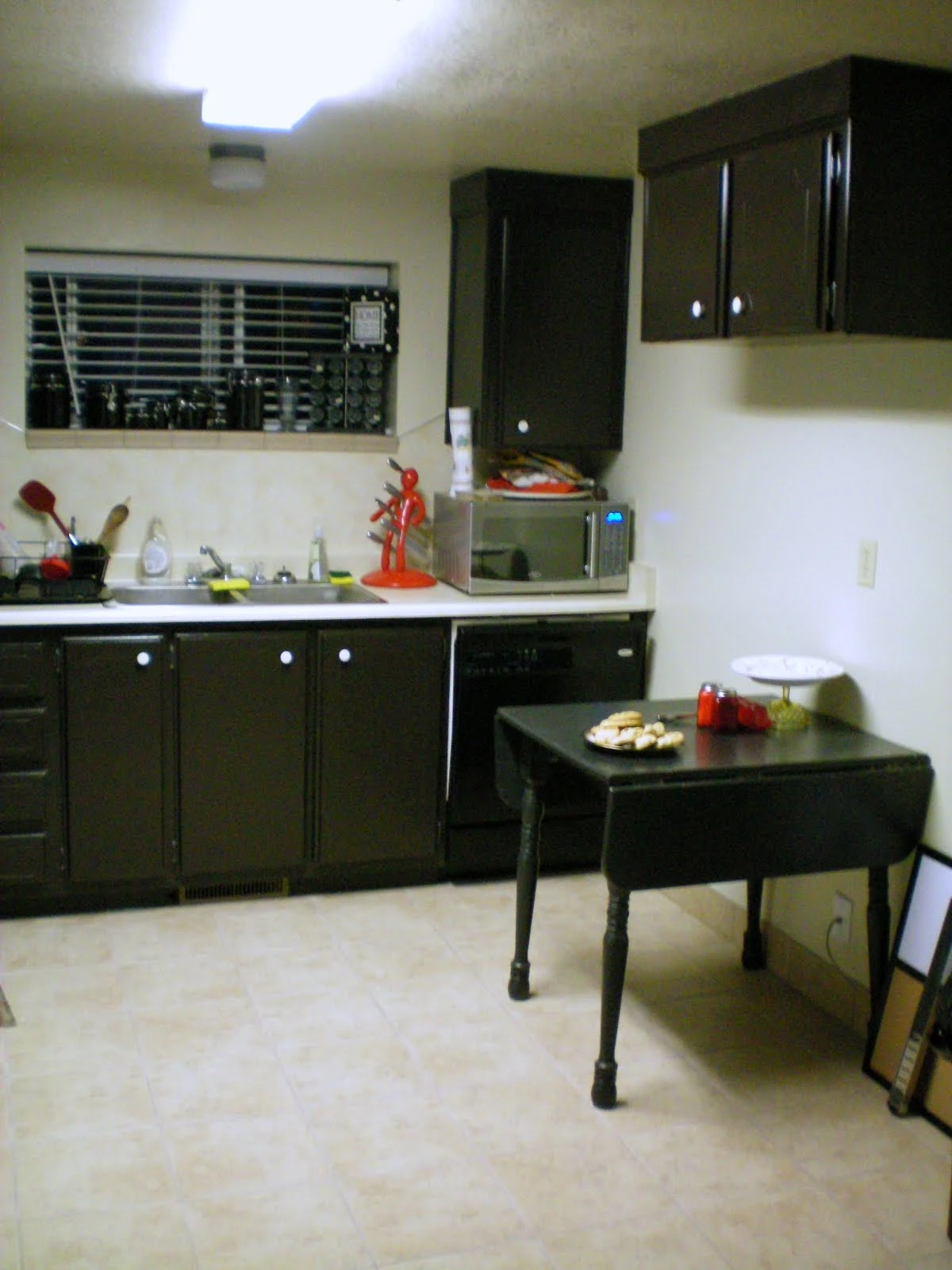 Kitchen with dark wood-finish cabinets, white tile floor, countertop appliances including a microwave and knife holder near the window, and a small black drop-leaf table along the wall