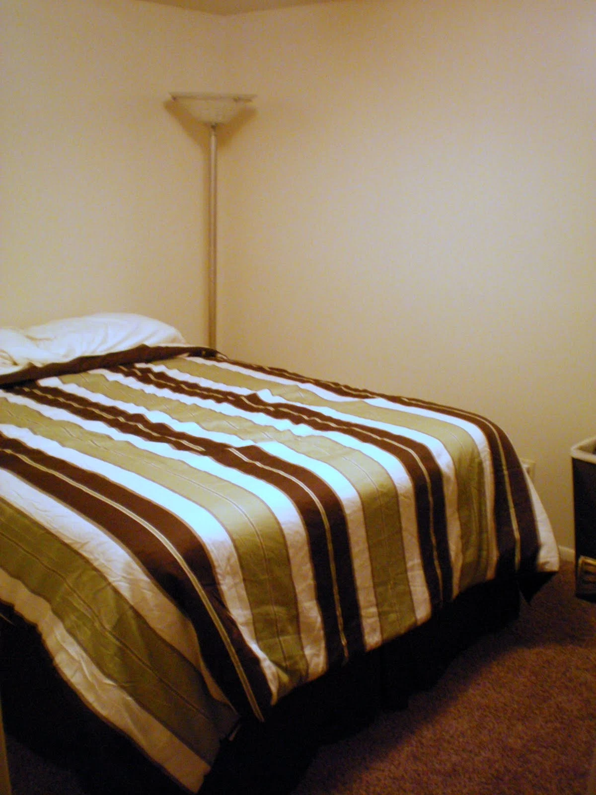 Bedroom corner showing the same striped bedspread, bare white walls, and a tall floor lamp standing beside the bed
