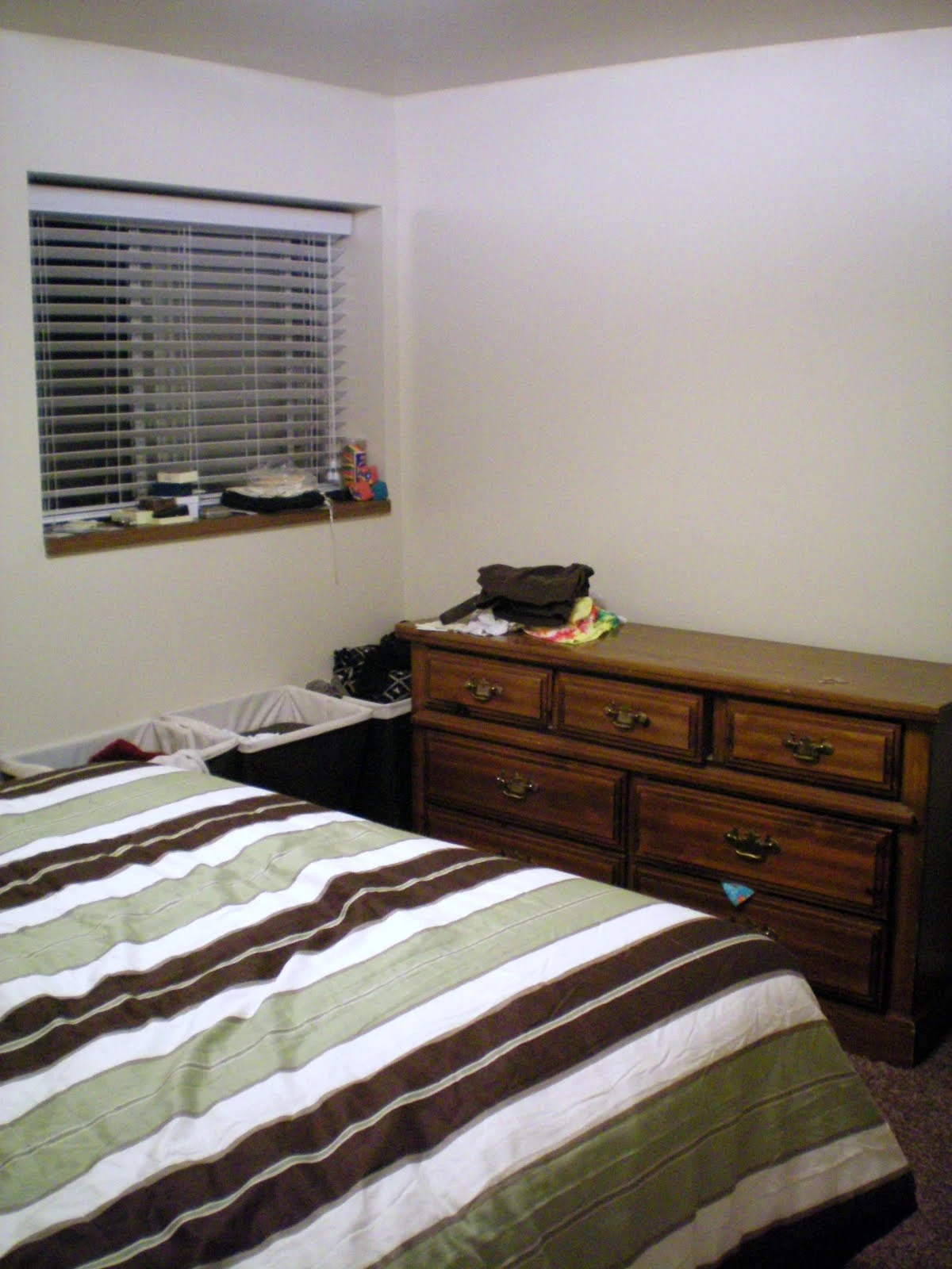 Bedroom with a striped brown, tan, green, and white bedspread, a large wooden multi-drawer dresser along the wall, and a window with horizontal blinds