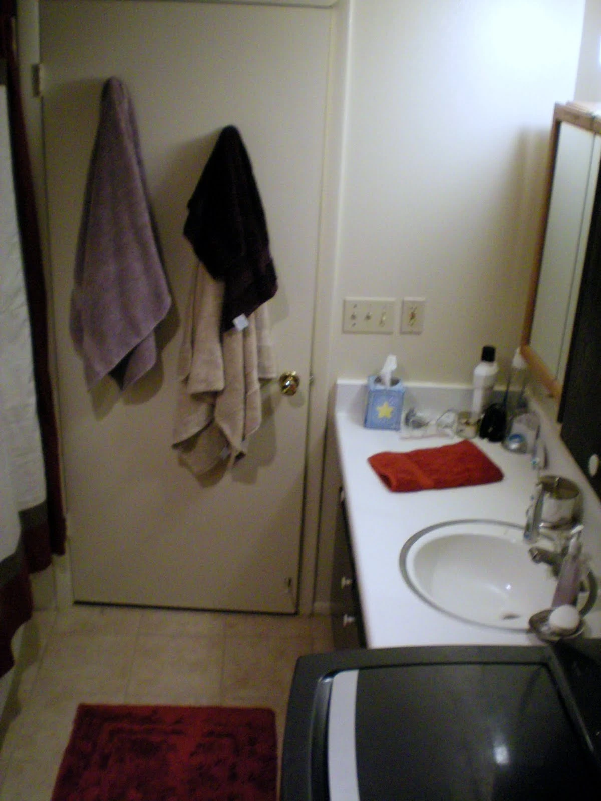 Small apartment bathroom with two towels hanging on the back of the door, a white single-basin sink and vanity with mirror, toiletries on the counter, a red bath mat, and a washer visible in the corner