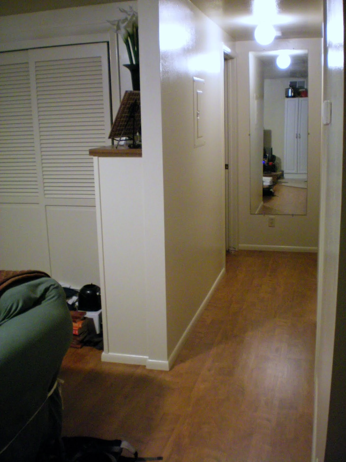 Narrow apartment entryway hallway with white louvered closet doors on the left, hardwood floors, and a full-length mirror at the far end reflecting the space
