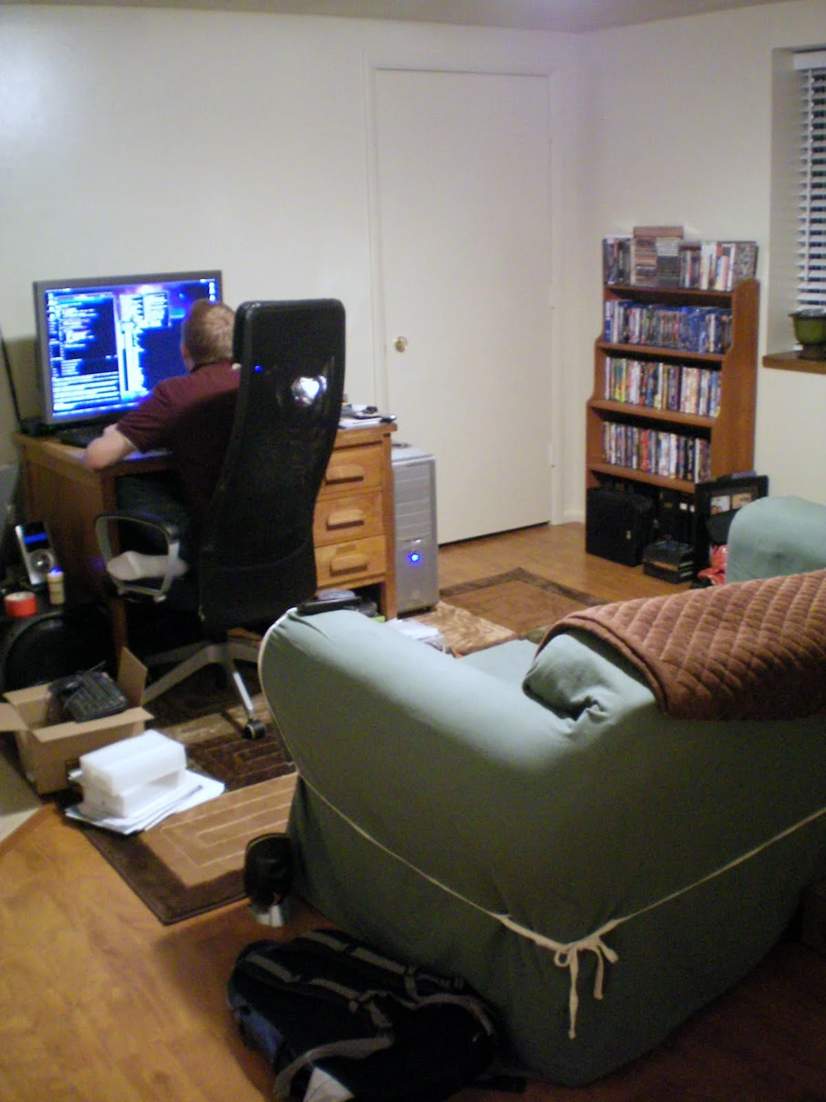 Jon seated at a computer desk in the living room, with a black office chair, tower PC, large monitor, and a tall bookshelf of DVDs along the far wall; a green armchair and area rug in the foreground