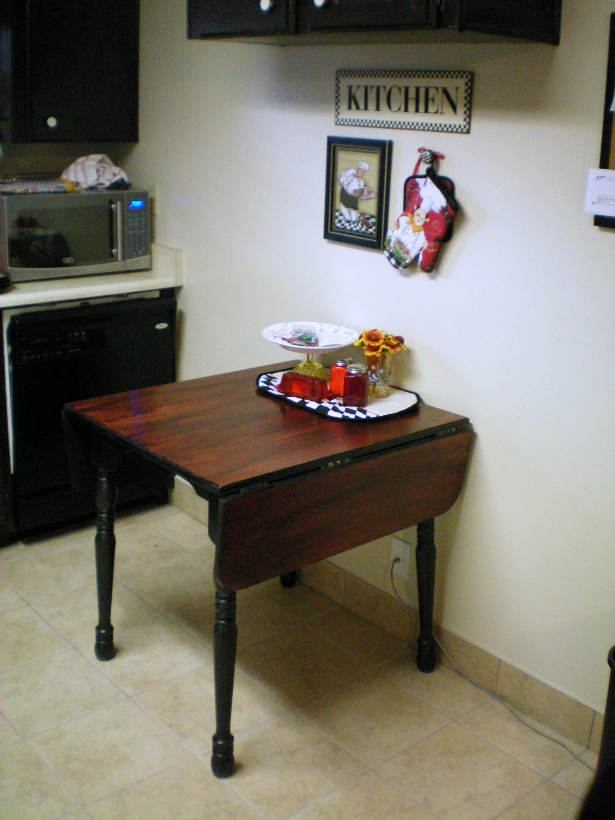 The finished kitchen table inside with a dark red stain and black turned legs, set in the kitchen corner with a "KITCHEN" sign and decorative rooster on the wall behind it