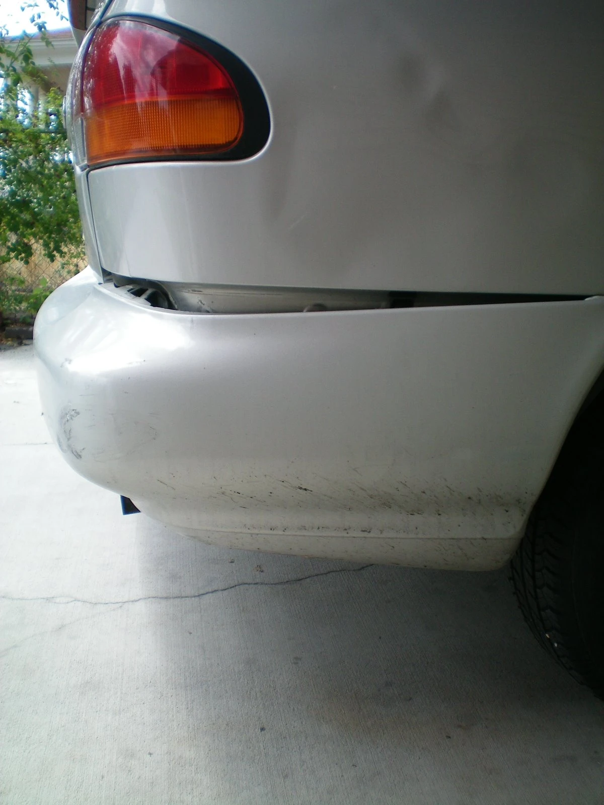 Close-up of the rear-left corner of the silver Nissan showing the bumper cracked and pushed inward, with the tail light visible above