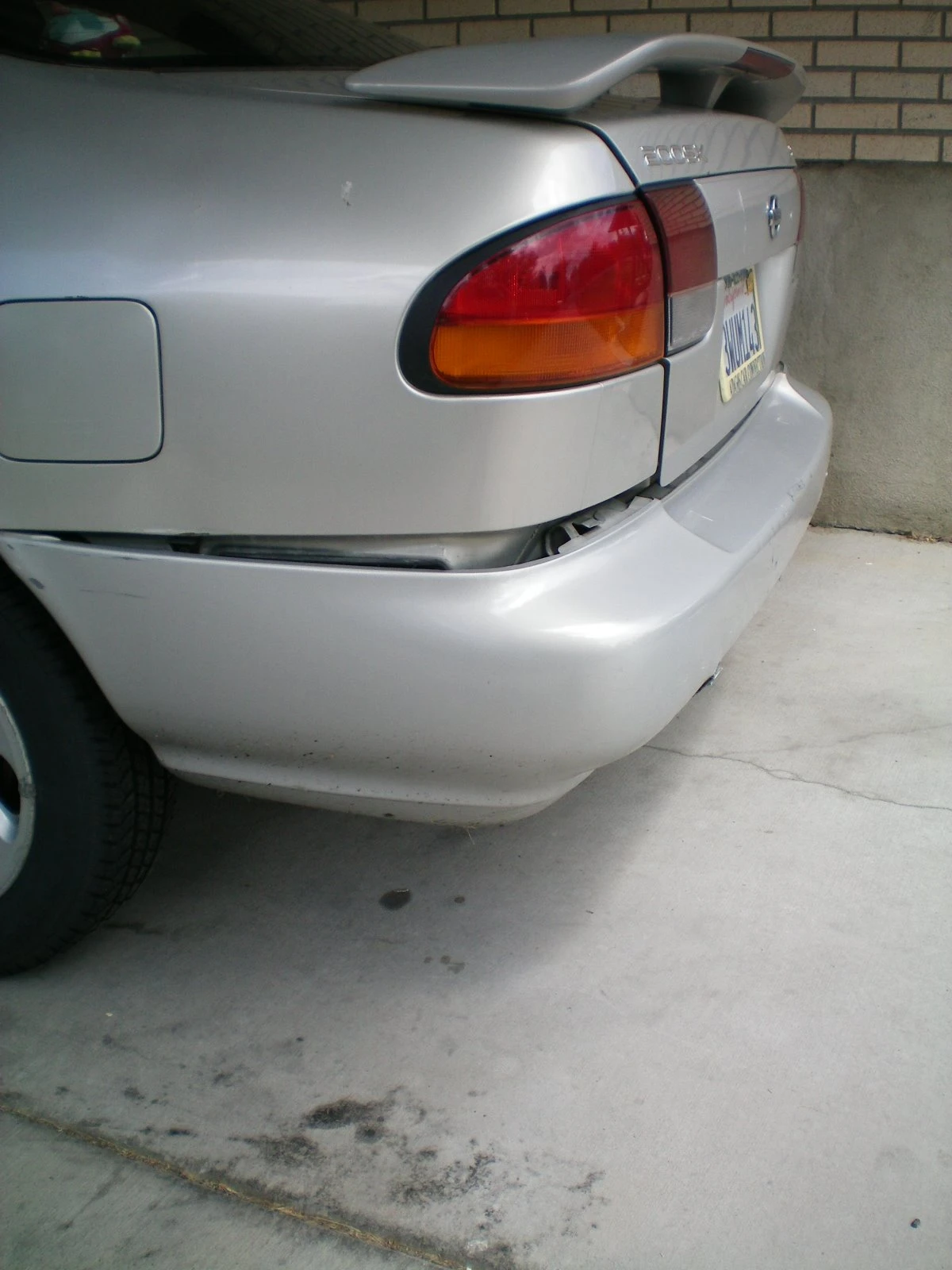 Rear of the silver Nissan showing the bumper pushed in and misaligned on the right side near the trunk, with a spoiler visible on top