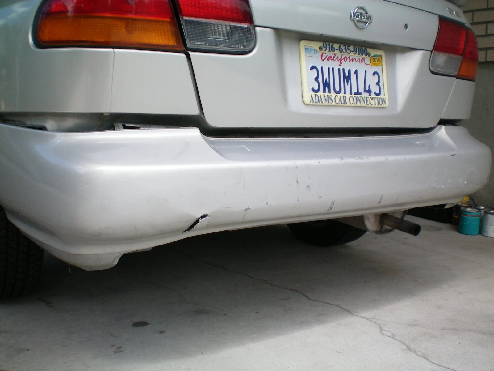 Rear of the silver Nissan with California plates showing scrapes and impact damage along the lower bumper with the exhaust pipe visible underneath