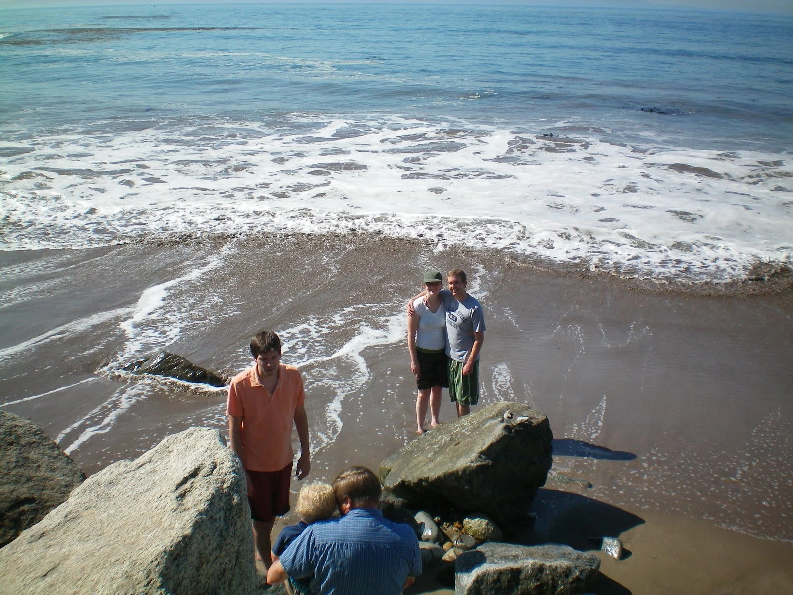 The family standing on wet sand at the beach with waves breaking behind them, viewed from above a rocky outcropping in the foreground