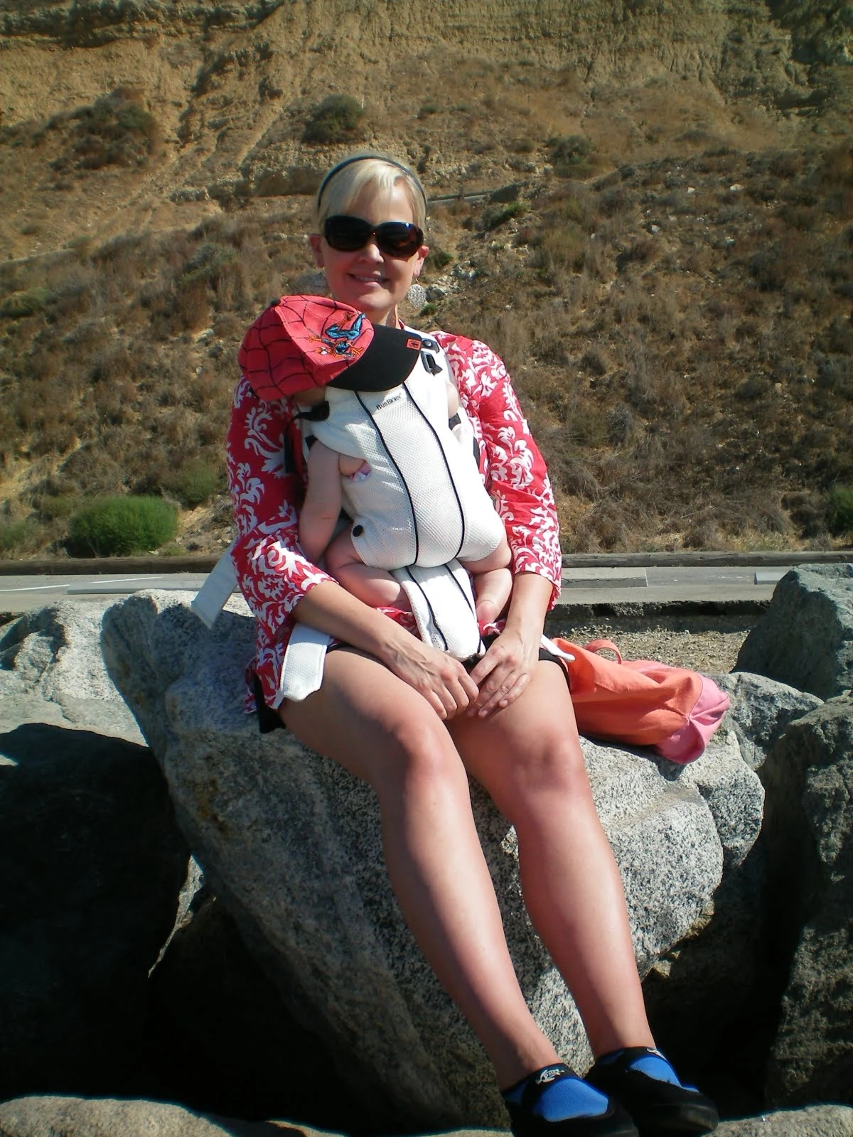 A woman in a red and white floral rash guard sitting on beach rocks holding a young baby, dry brown hills visible in the background