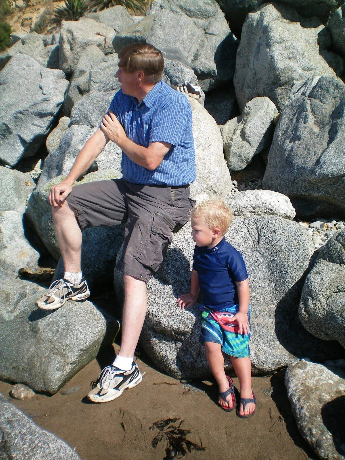 An older man in a blue shirt sitting on beach rocks while a young blonde toddler boy in a blue shirt and colorful swim shorts stands close beside him