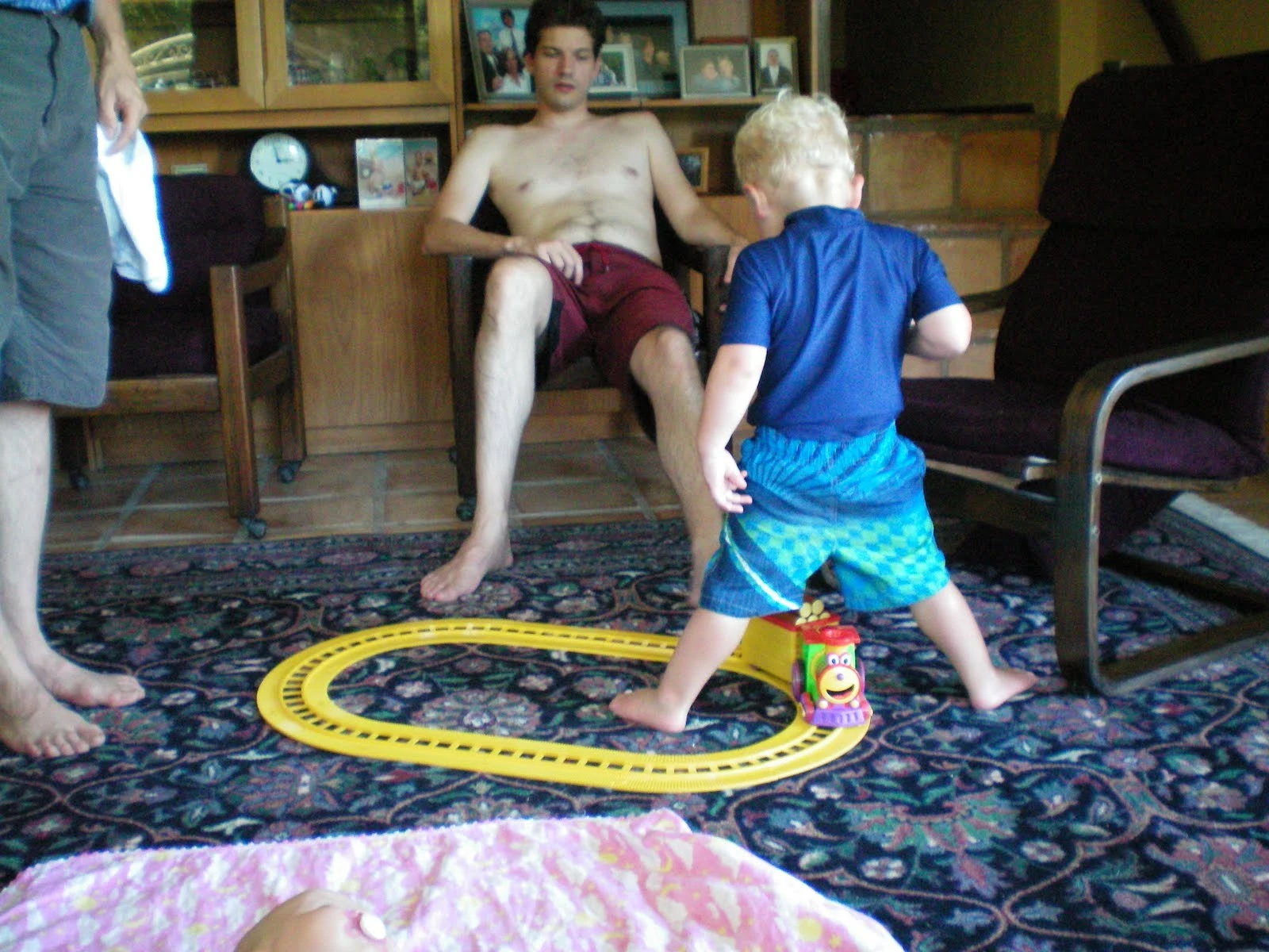 A shirtless man in red swim trunks relaxing in a living room chair while a blonde toddler plays with a yellow toy train track on a patterned rug, a baby lying on the floor in the foreground