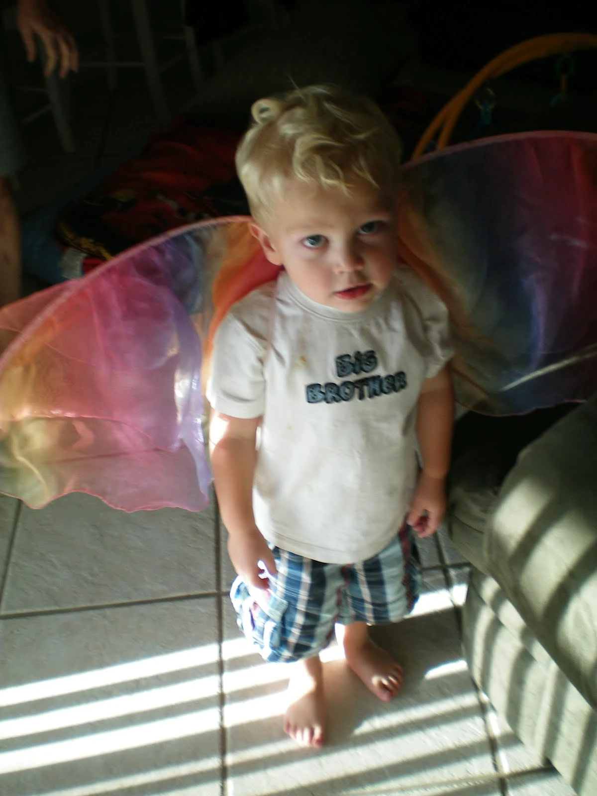 Charlie, a young blonde toddler, wearing a white "Big Brother" shirt and plaid shorts, standing barefoot on tile and looking up at the camera