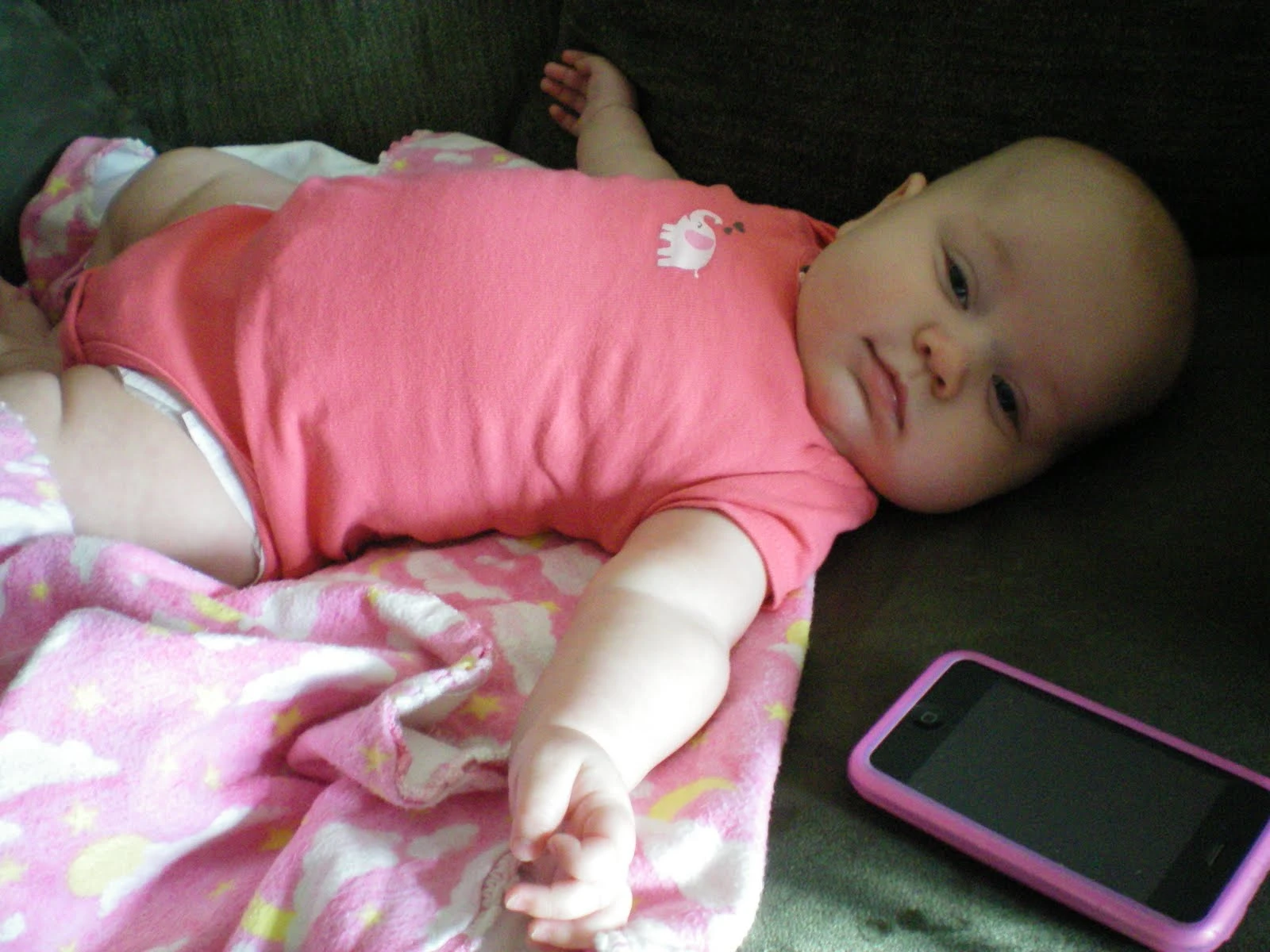 Baby Jillian in a pink onesie lying on a dark couch, reaching one arm out toward the camera, a pink phone case beside her