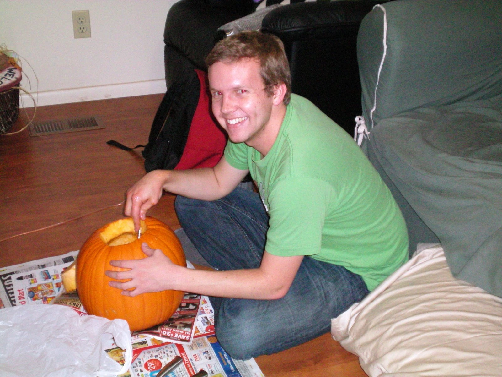 Jon in a green shirt kneeling on newspaper-covered floor, smiling while scooping out a pumpkin