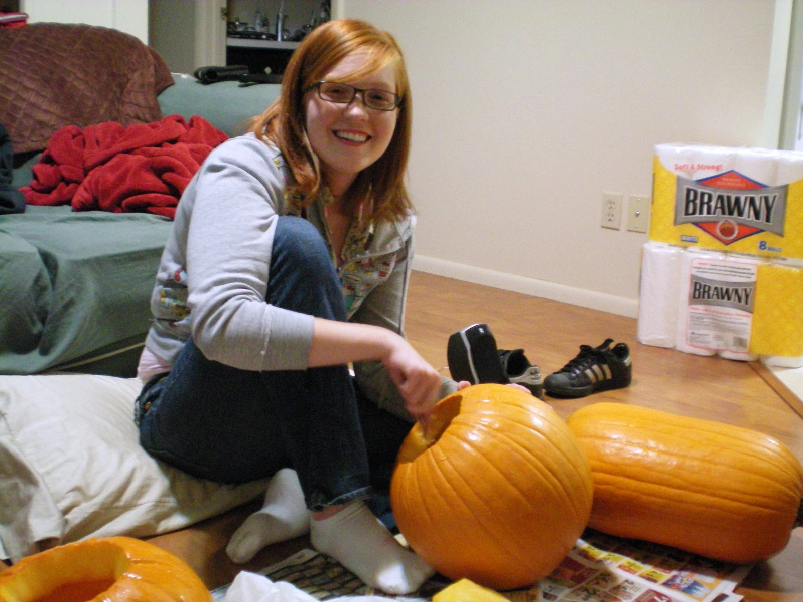 Danielle in a grey hoodie kneeling on the floor carving one of three pumpkins laid out on newspaper, a roll of Brawny paper towels nearby