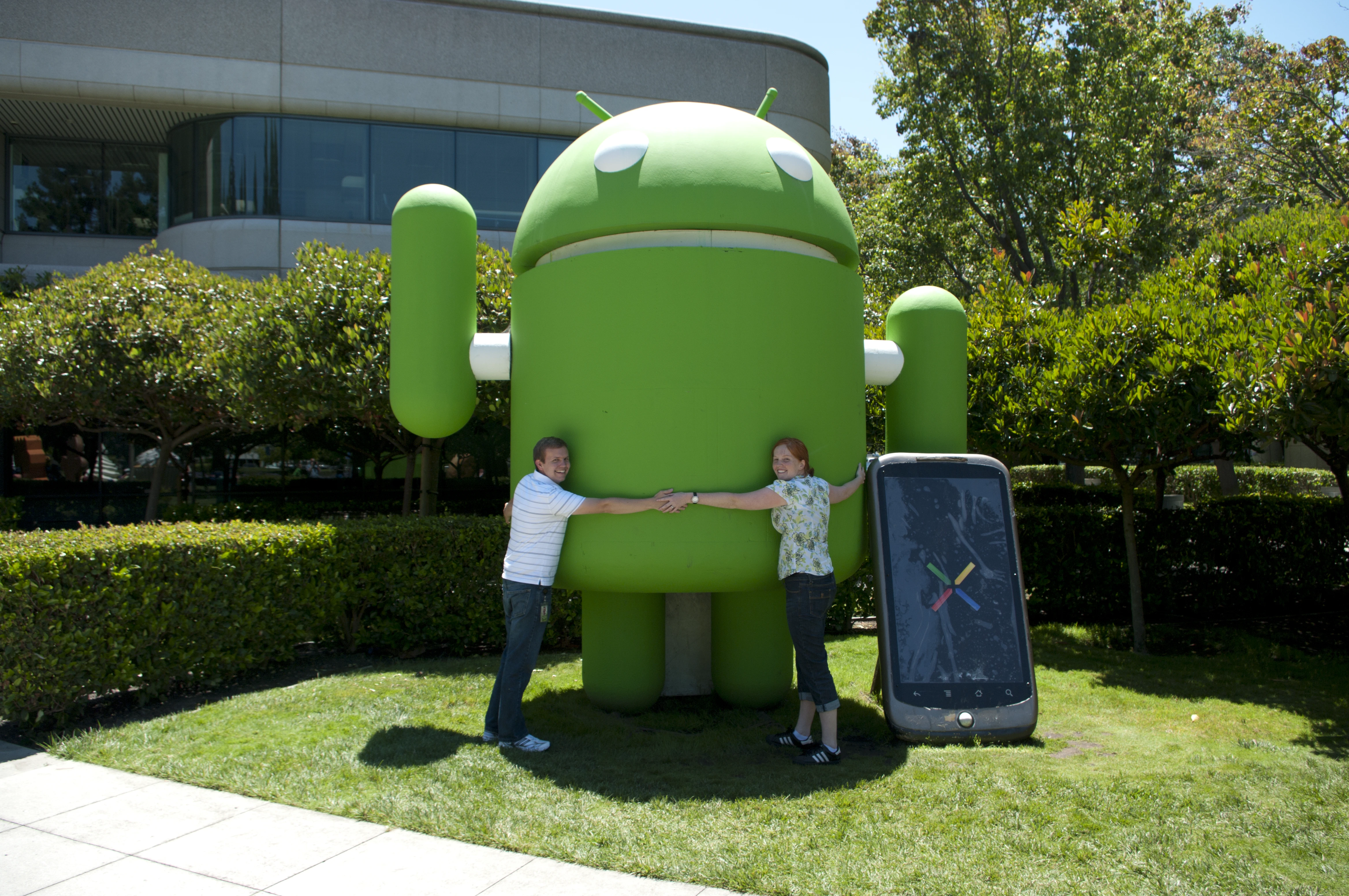 Jon and Danielle standing on either side of the giant green Android robot statue on the Google campus lawn, each holding one of its hands, with a large Nexus One phone sculpture beside it