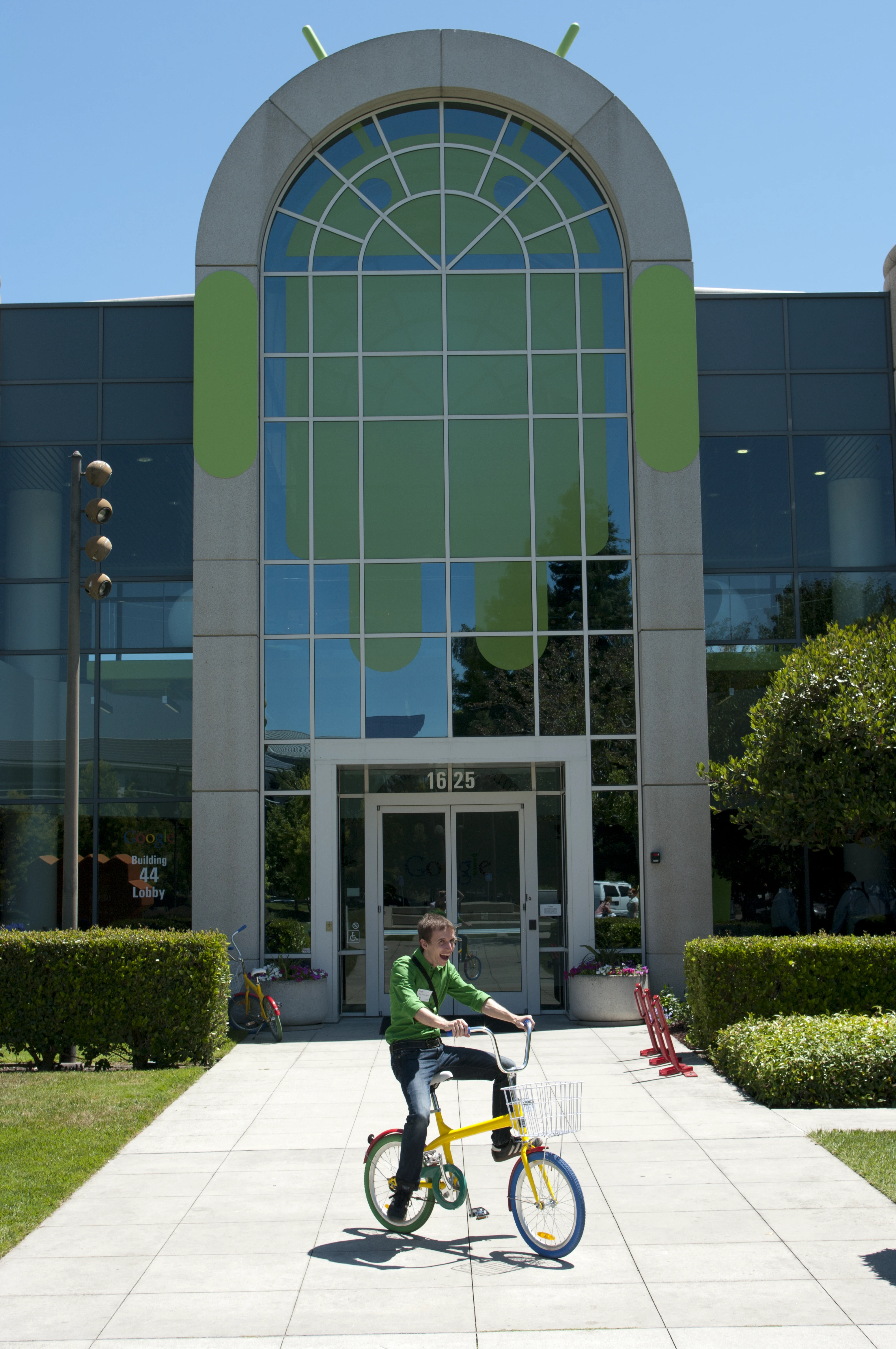 Ryan riding a yellow, blue, and red Google bike on a path in front of the Android building (Building 16125), which features a large arched Android logo window