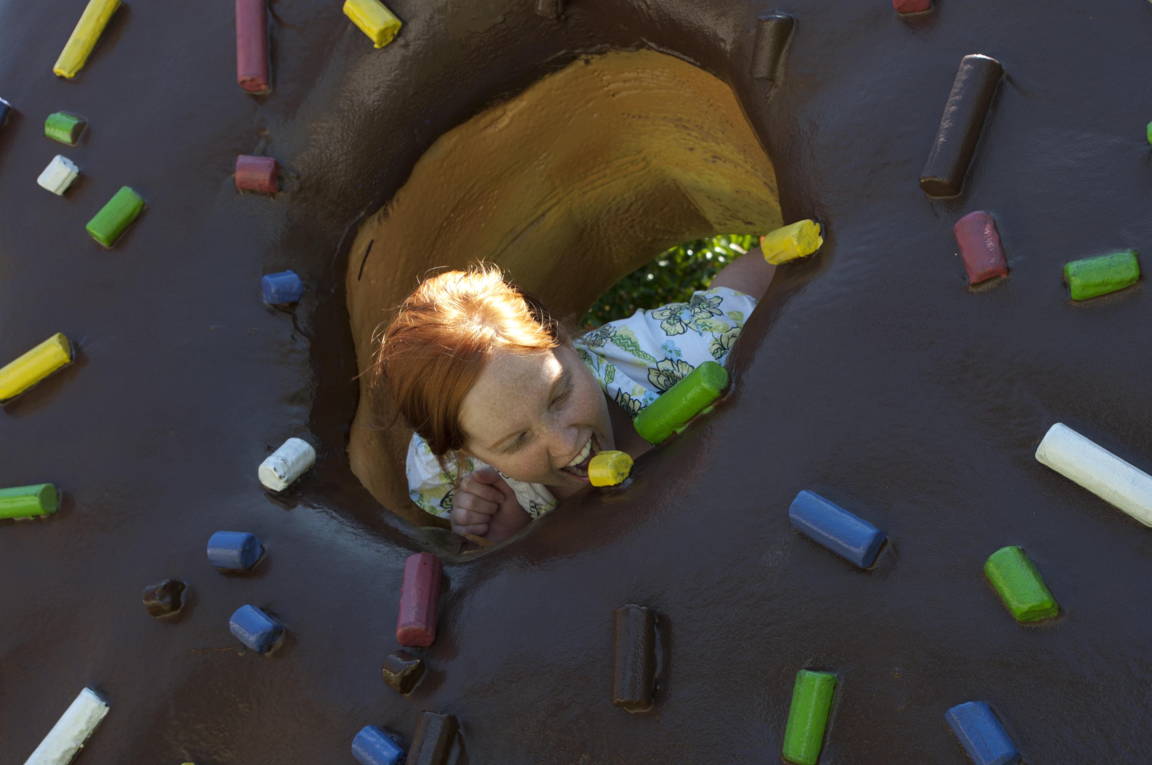 Danielle peeking through the center hole of the giant Android Donut sculpture, photographed from above, surrounded by colorful candy-shaped sprinkles in red, yellow, green, and blue