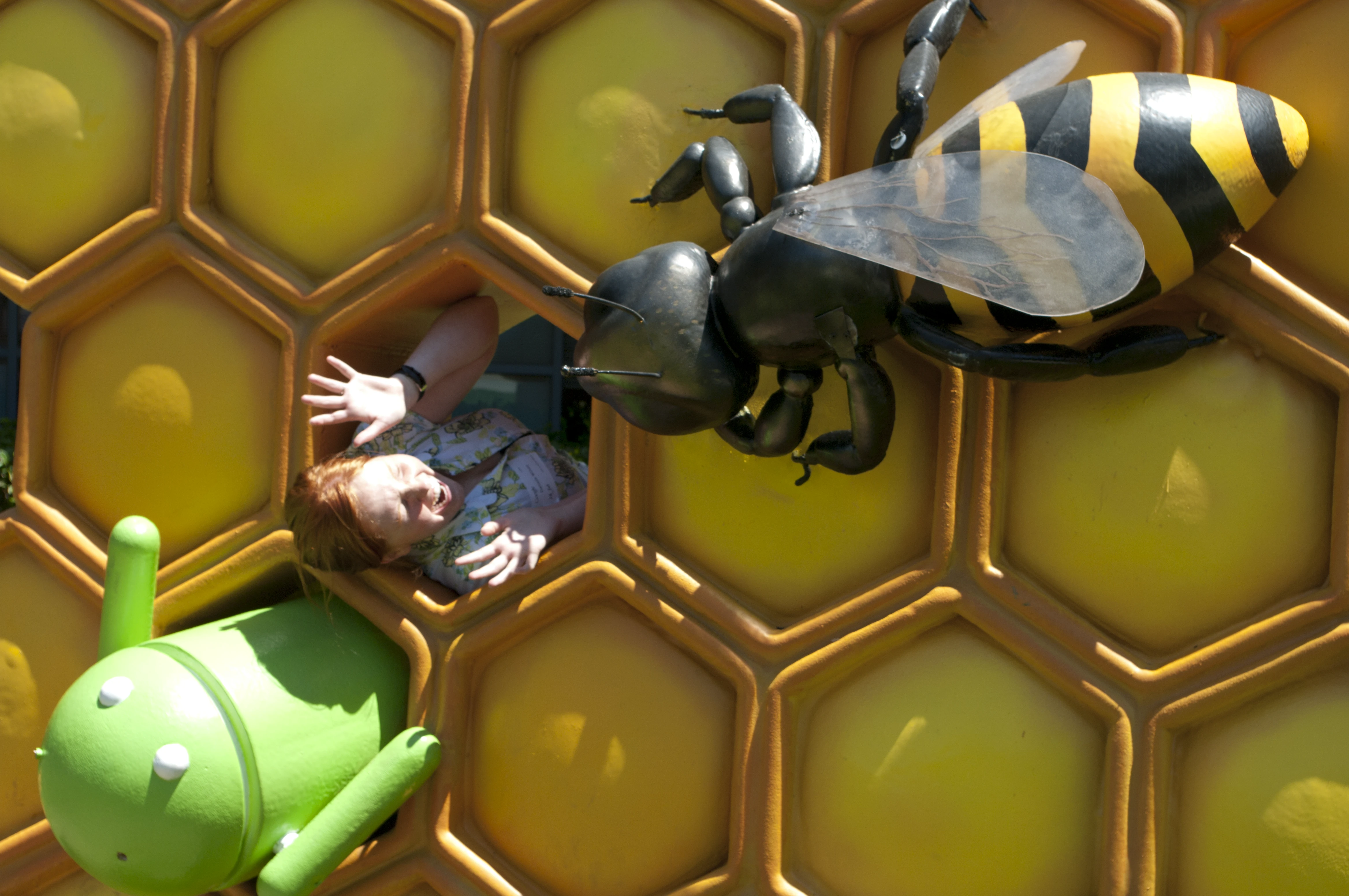 Danielle lying on a giant honeycomb sculpture being "attacked" by a large bee statue, arms raised in mock alarm, with a small green Android figurine in the lower left corner