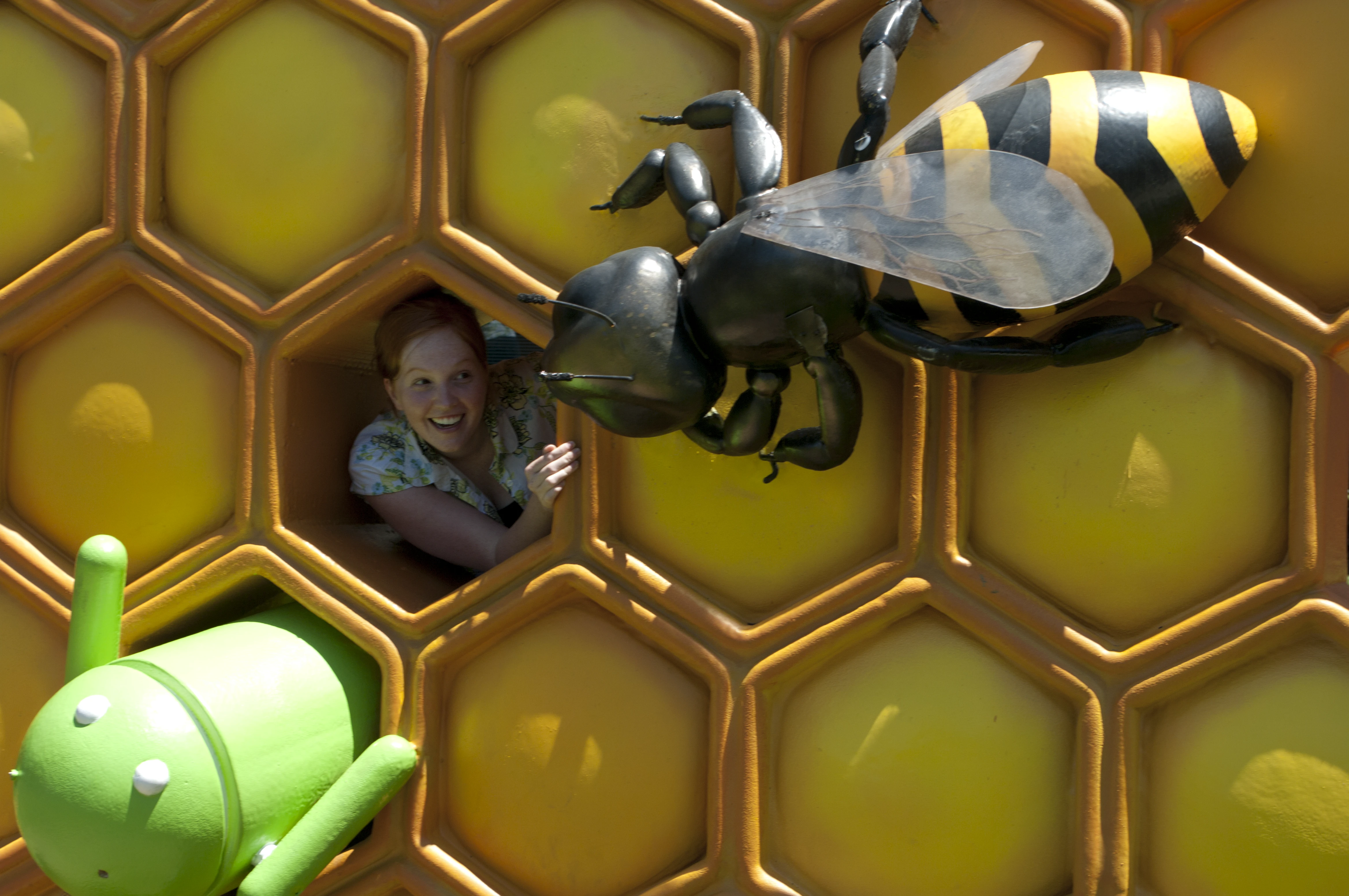 Danielle smiling from inside a giant honeycomb cell with the large bee sculpture looming in her face, and a small green Android figurine in the lower left corner