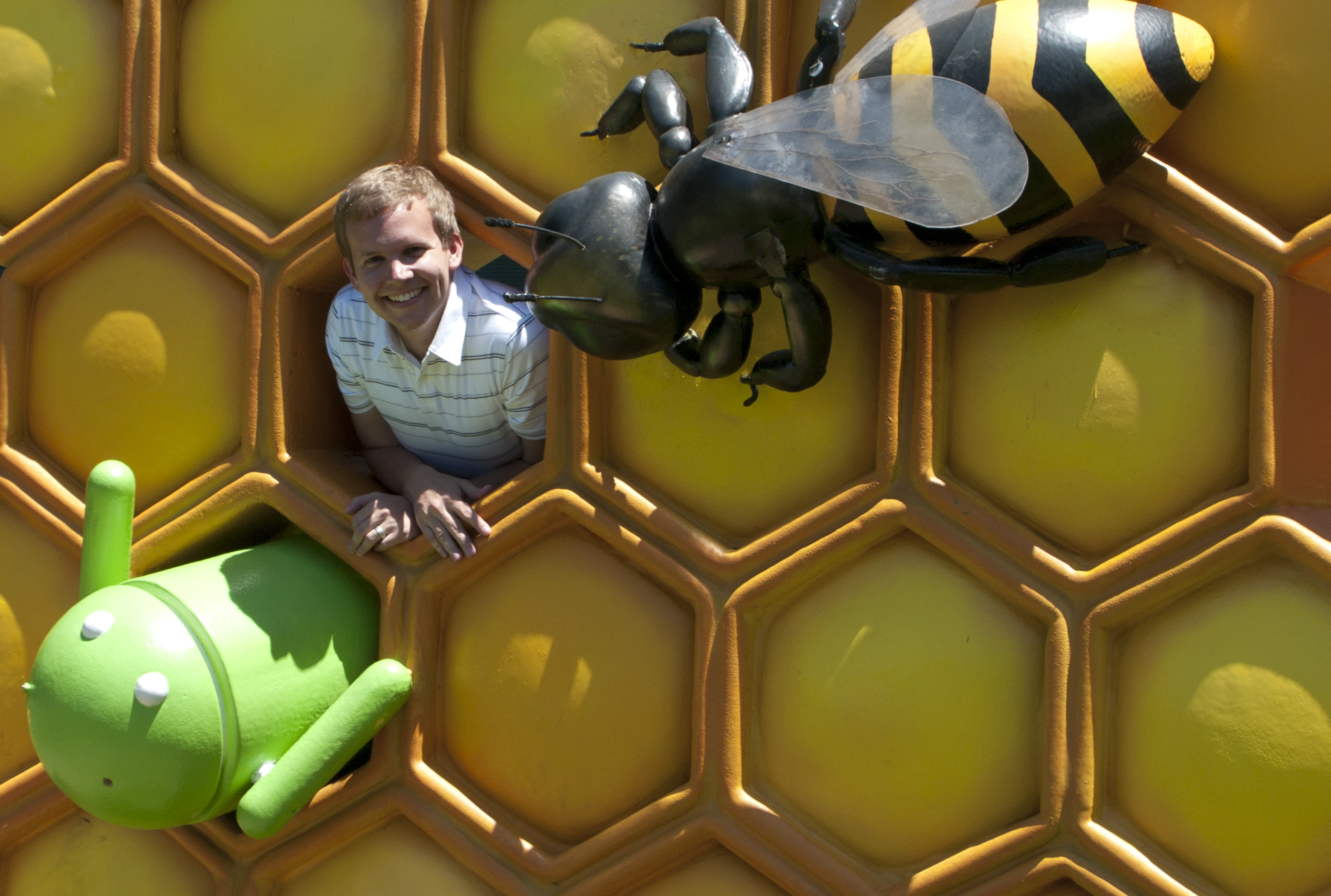 Jon smiling while peeking out from inside a giant honeycomb cell, with the large bee sculpture above him and a small green Android figurine beside him