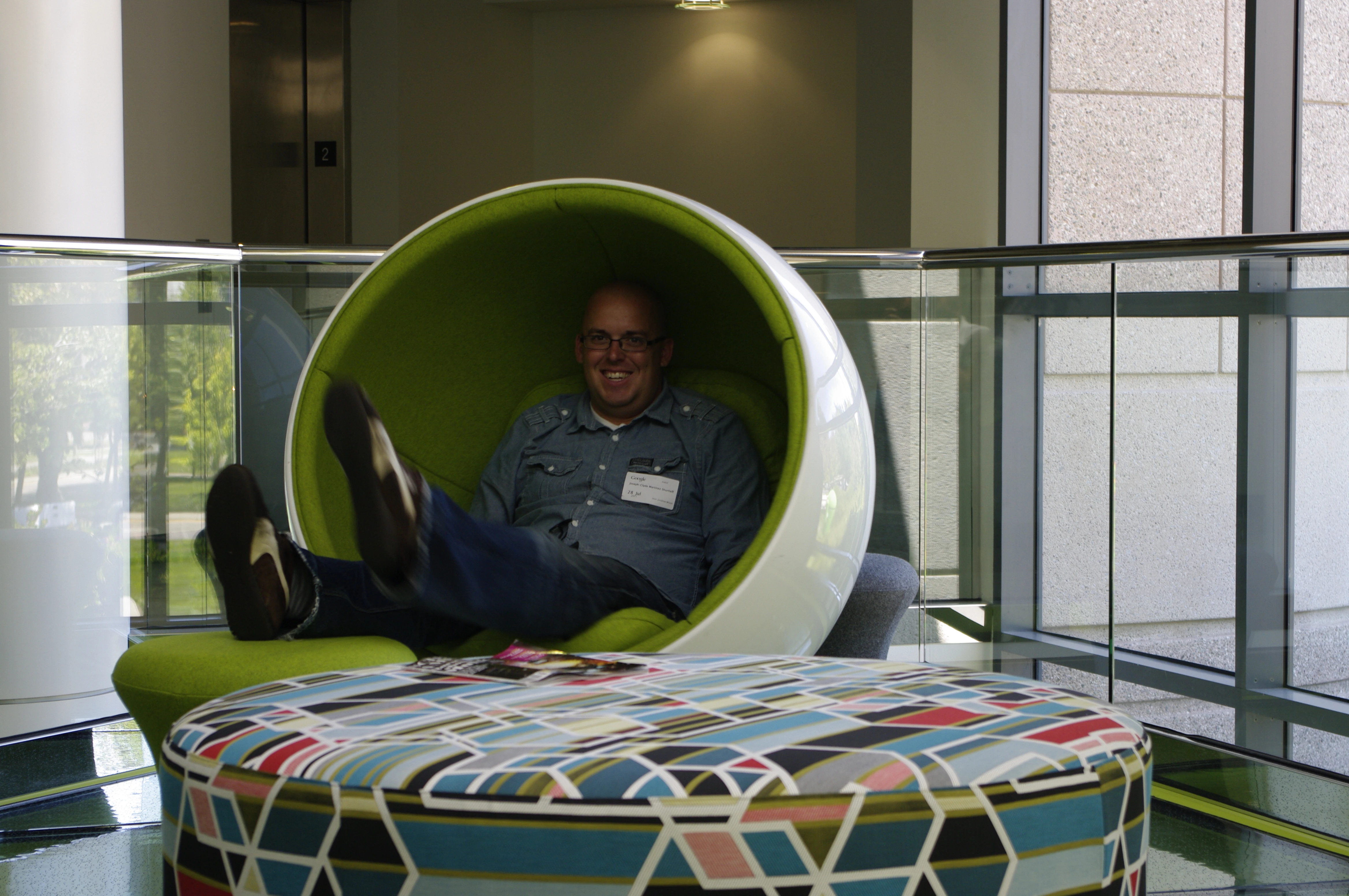 Joe smiling while lounging in a large green egg-shaped pod chair inside a Google building, feet propped up, with a colorful geometric-patterned ottoman in front