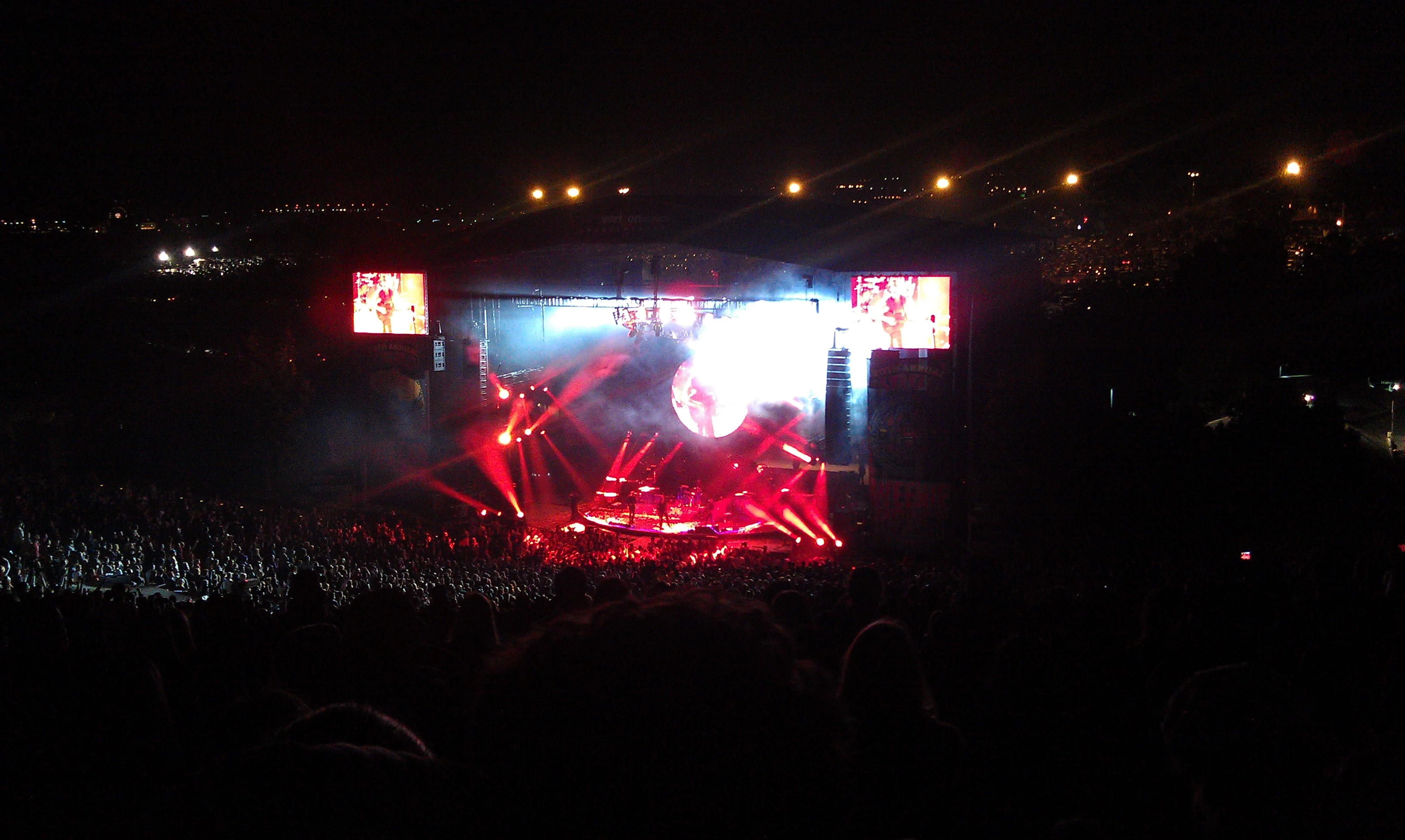Nighttime concert with dramatic red and pink stage lighting, a large glowing sphere at center stage, banks of spotlights fanning out over a huge silhouetted crowd