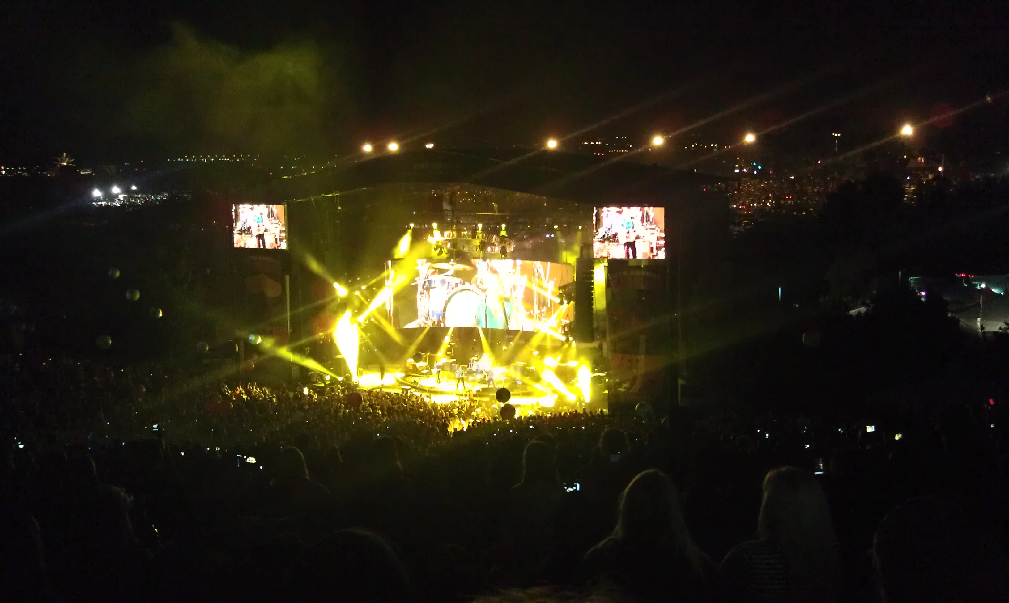 Nighttime concert with bright golden-yellow lighting and smoke effects, large LED screens on both sides of the stage showing performers, crowd visible in the foreground with phones raised