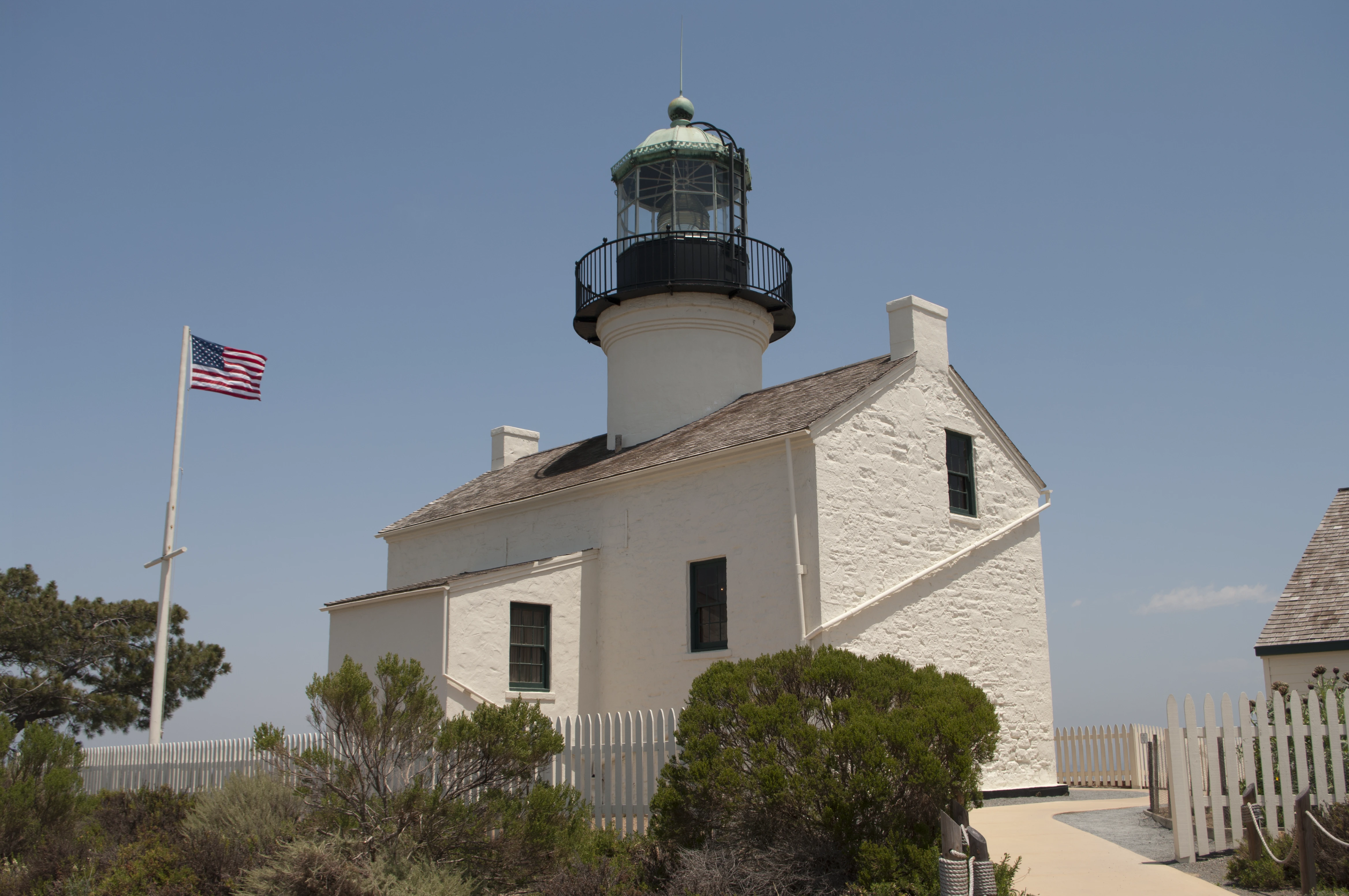 The Old Point Loma Lighthouse, a white stone building with a circular tower topped by a green dome, an American flag on a tall pole to the left, surrounded by scrubby brush and a white picket fence
