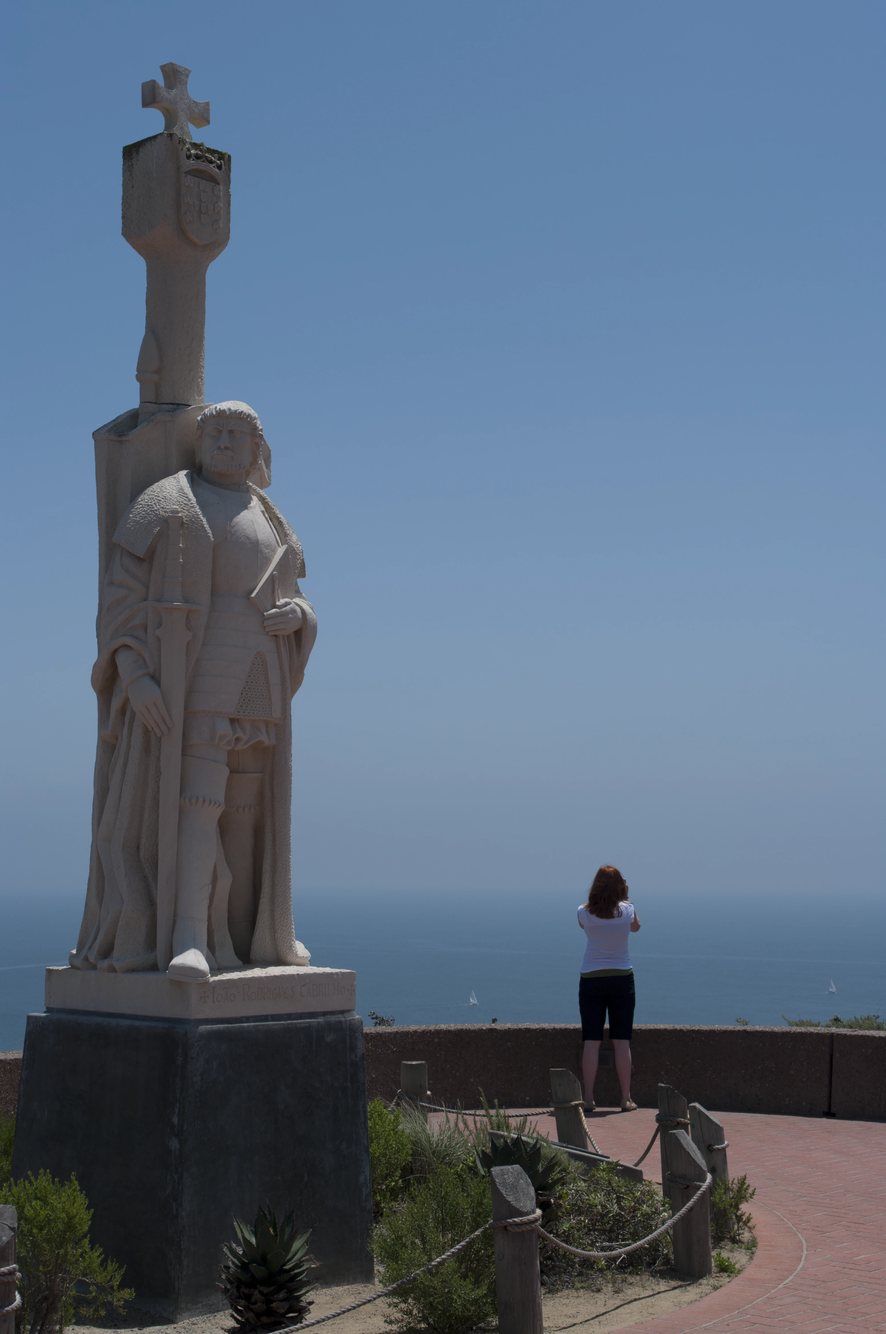 Danielle standing with her back to the camera next to the Cabrillo statue, her red hair visible, looking out over the ocean in bright sunlight